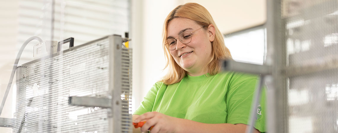 Eine Person mit blonden Haaren in einem grünen Shirt arbeitet an einem technischen Gerät mit einem Werkzeug in der Hand.