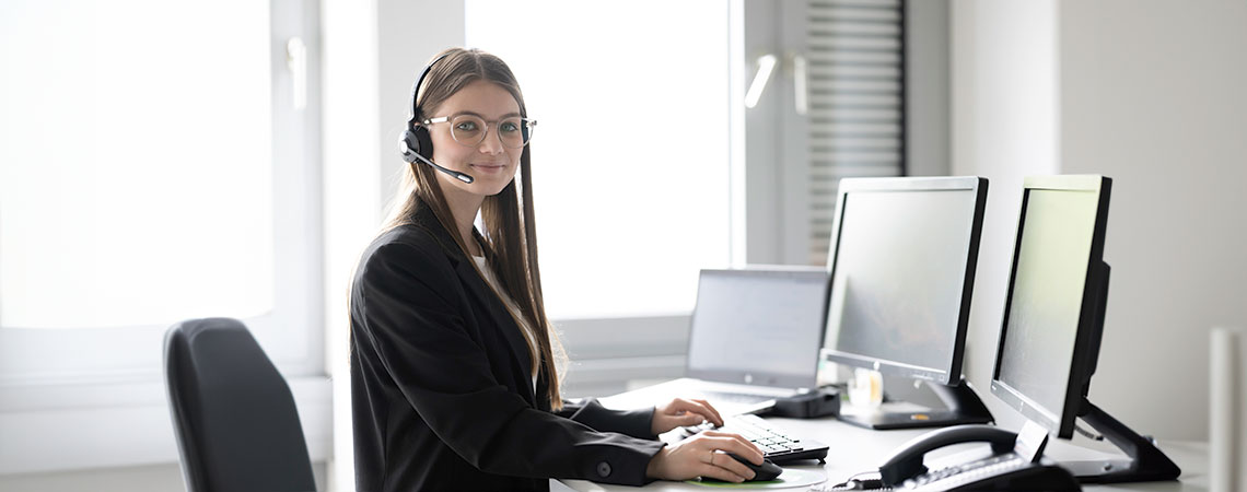 Junge Frau mit Headset sitzt an einem Schreibtisch mit mehreren Monitoren und arbeitet am Computer.