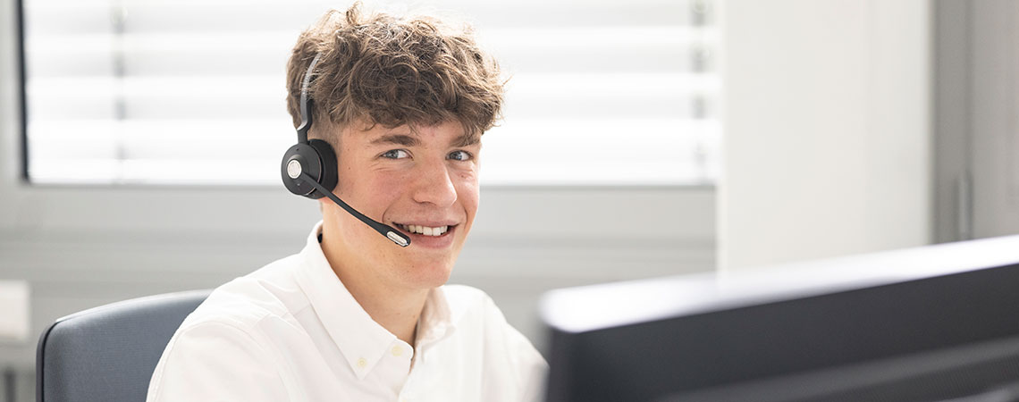 Junger Mann mit Headset, lächelt, sitzt am Schreibtisch vor einem Computerbildschirm, helle Büroumgebung.