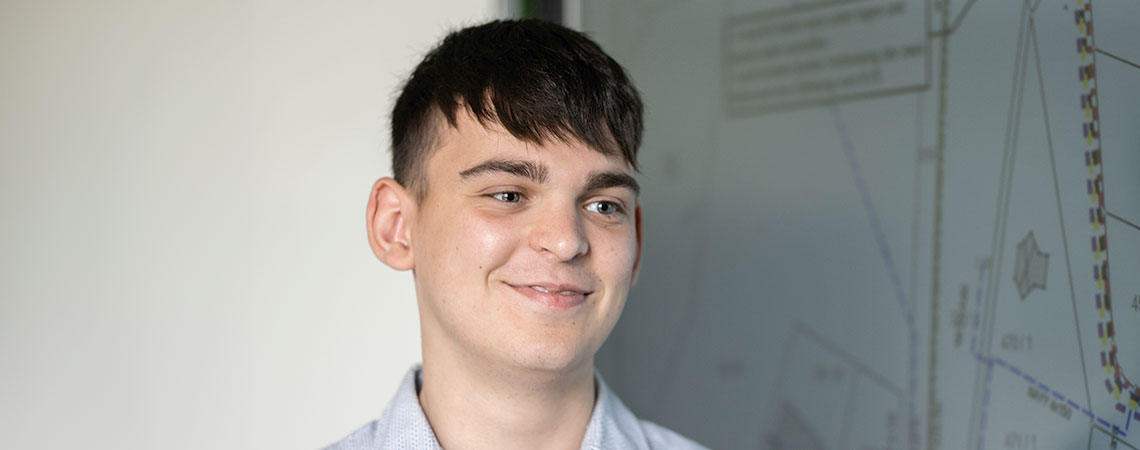Ein junger Mann mit kurzem, dunklem Haar lächelt vor einer Wand mit Plänen oder Zeichnungen.