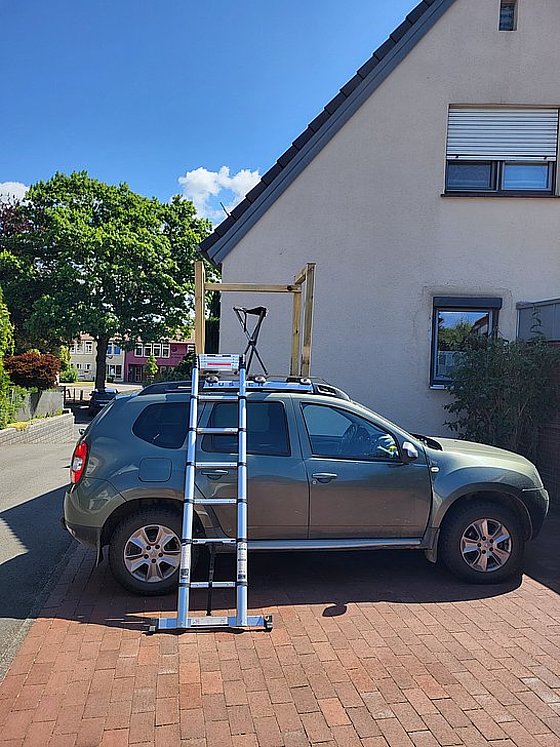 Grünes Auto mit einer Aluminiumleiter, die auf das Dach führt, vor einem Wohnhaus und Bäumen im Hintergrund.
