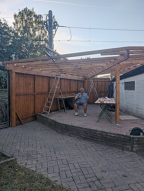 Mann sitzt unter einer Holzkonstruktion und spielt Gitarre, umgeben von einer Gartenlandschaft und Arbeitsmaterial.