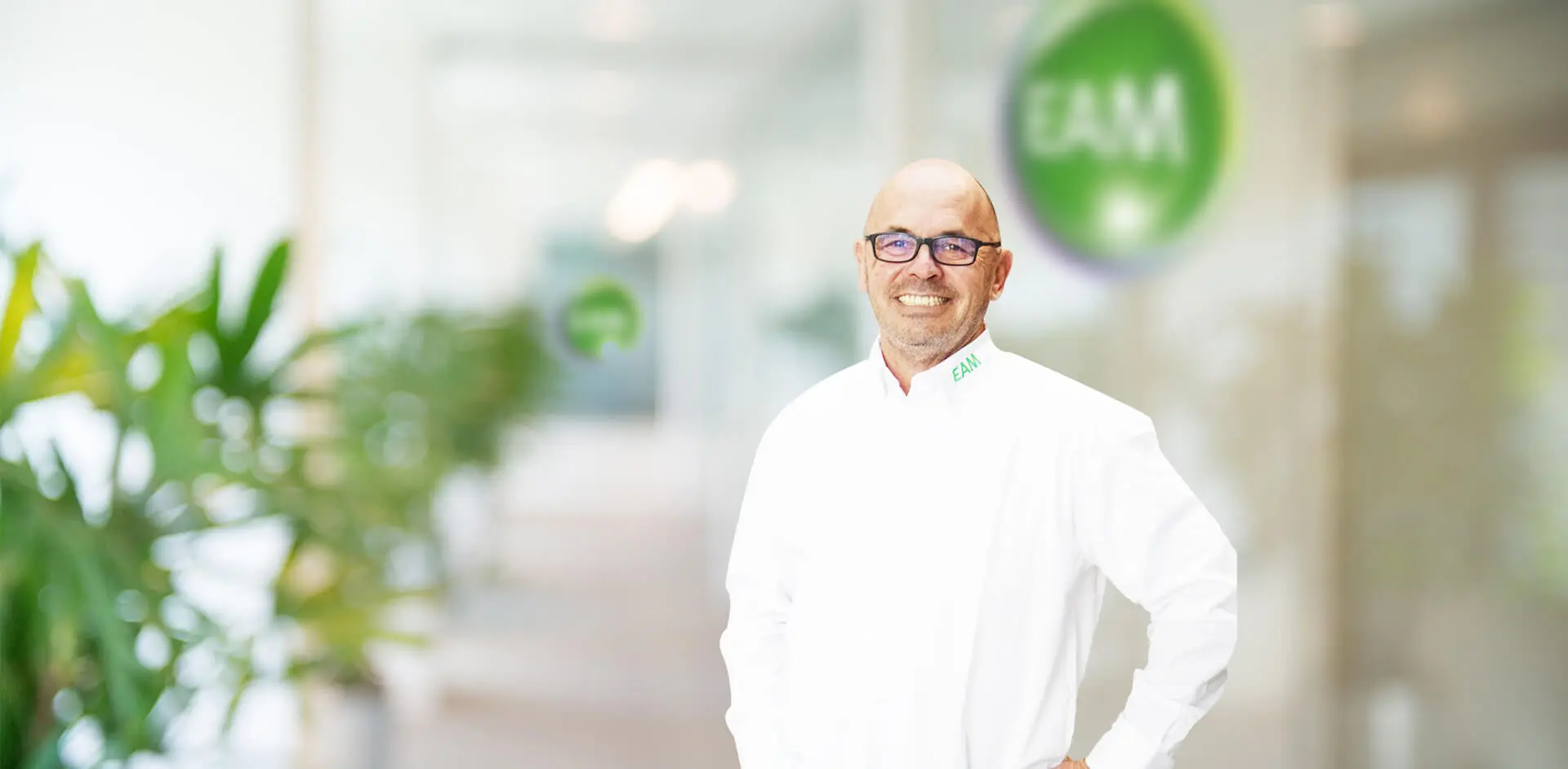 Ein lachender Mann mit Brille und Bart steht vor einem EAM-Logo in einem Büro.