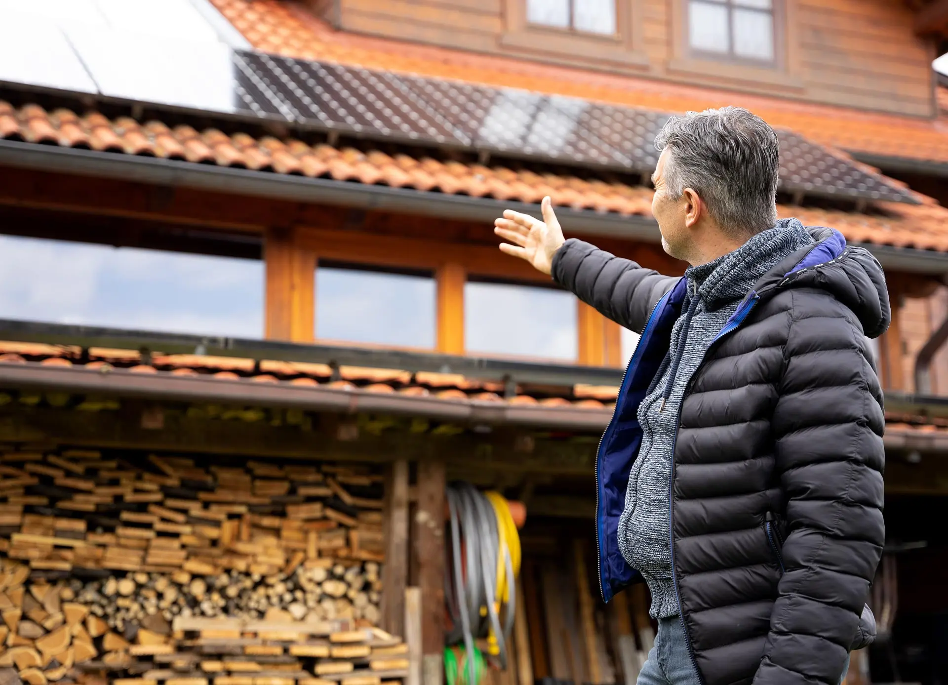 Mann steht vor einem Holzhaus und zeigt auf die Solarpanels auf dem Dach. Holzstapel und Werkzeuge im Hintergrund sichtbar.