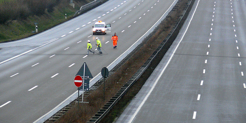 Drei Arbeiter in Warnkleidung führen Reparaturen auf einer leeren Straße durch, während ein Fahrzeug mit Warnlicht steht.
