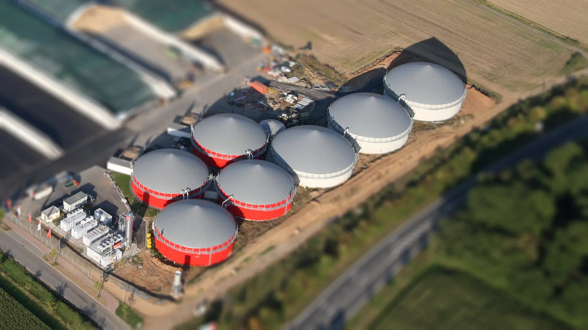  Luftbild einer Biogasanlage mit sieben runden Fermentern, teils rot, teils weiß, inmitten landwirtschaftlicher Felder.