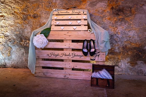 Holzregal mit dem Schriftzug "High Heels Garage", schwarzer Schuh, weiße Blume und transparenten Accessoires.