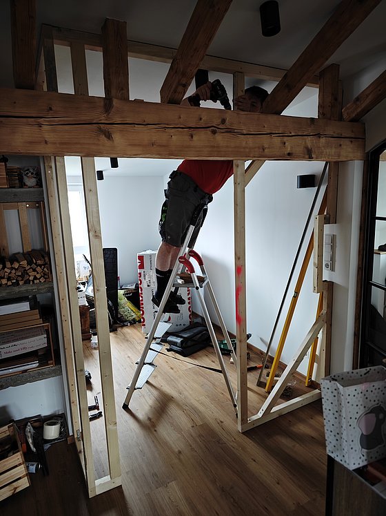 Mann auf einer Leiter bei der Montage einer Holzstruktur im Innenraum, umgeben von Werkzeug und Materialien.