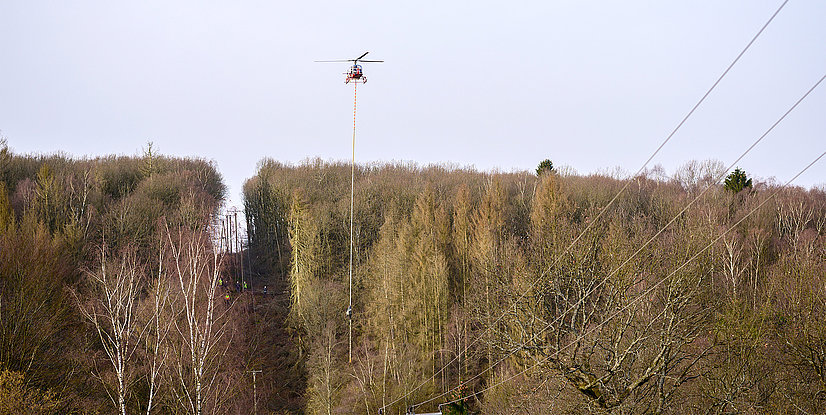 An einem Helikopter hängt eine große Säge, der Helikopter fliegt über einen Wald und bahnt mit der Säge einer Stromtrasse den Weg. 