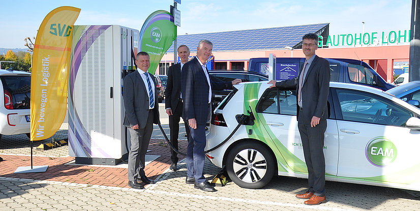 Vier Männer stehen neben einem Ladestellplatz für Elektroautos mit Werbebannern im Hintergrund. Ein Auto wird gerade geladen.