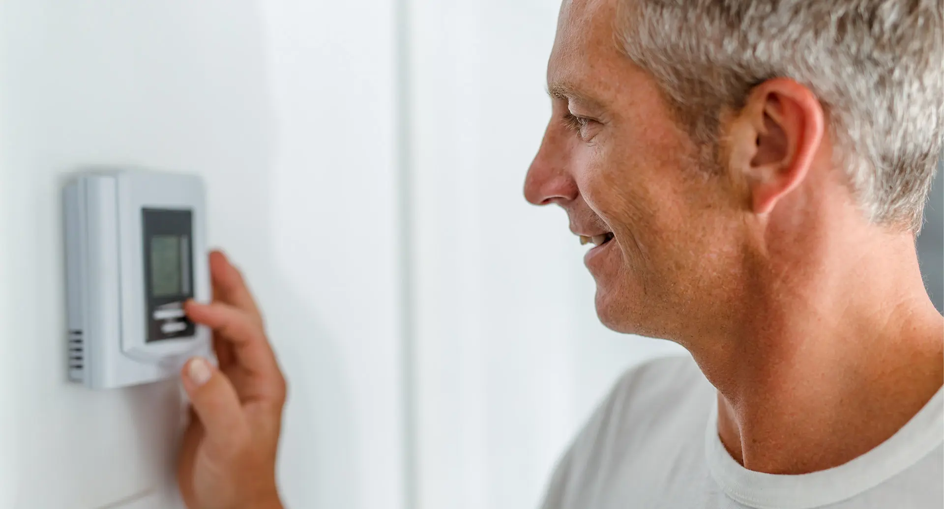 Mann mit grauen Haaren, der einen Thermostat an der Wand berührt und lächelt.