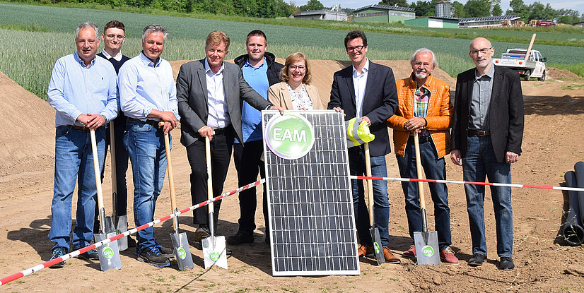 Gruppenfoto von Personen bei der Eröffnung eines Solarprojekts, mit Solarpanel und Schaufeln auf einem Feld.