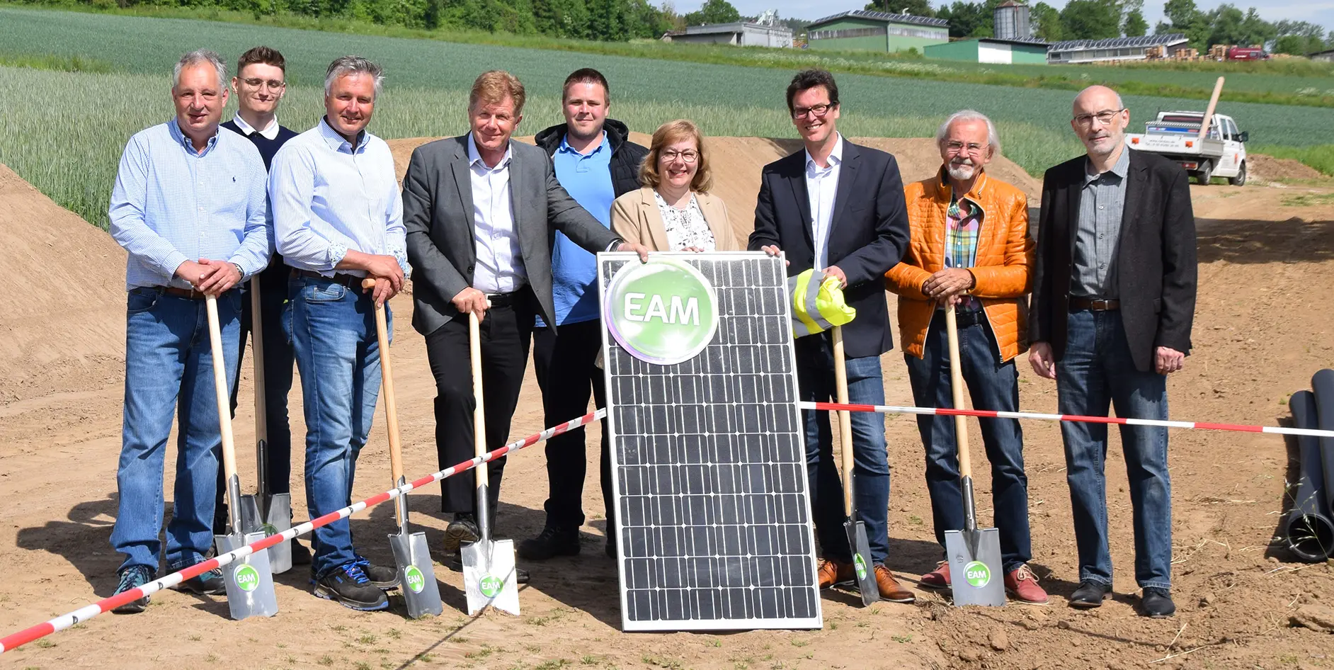 Gruppenfoto von Personen bei der Eröffnung eines Solarprojekts, mit Solarpanel und Schaufeln auf einem Feld.