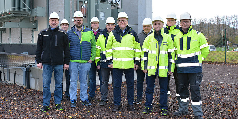 Gruppenschuss von Männern in Warnkleidung und Helmen vor technischen Anlagen, im Freien auf einem Schotterplatz.