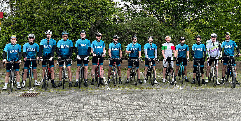 Gruppe von Radfahrern in blauer Teamsportkleidung, stehend neben ihren Fahrrädern, lächelnd in einer natürlichen Umgebung.