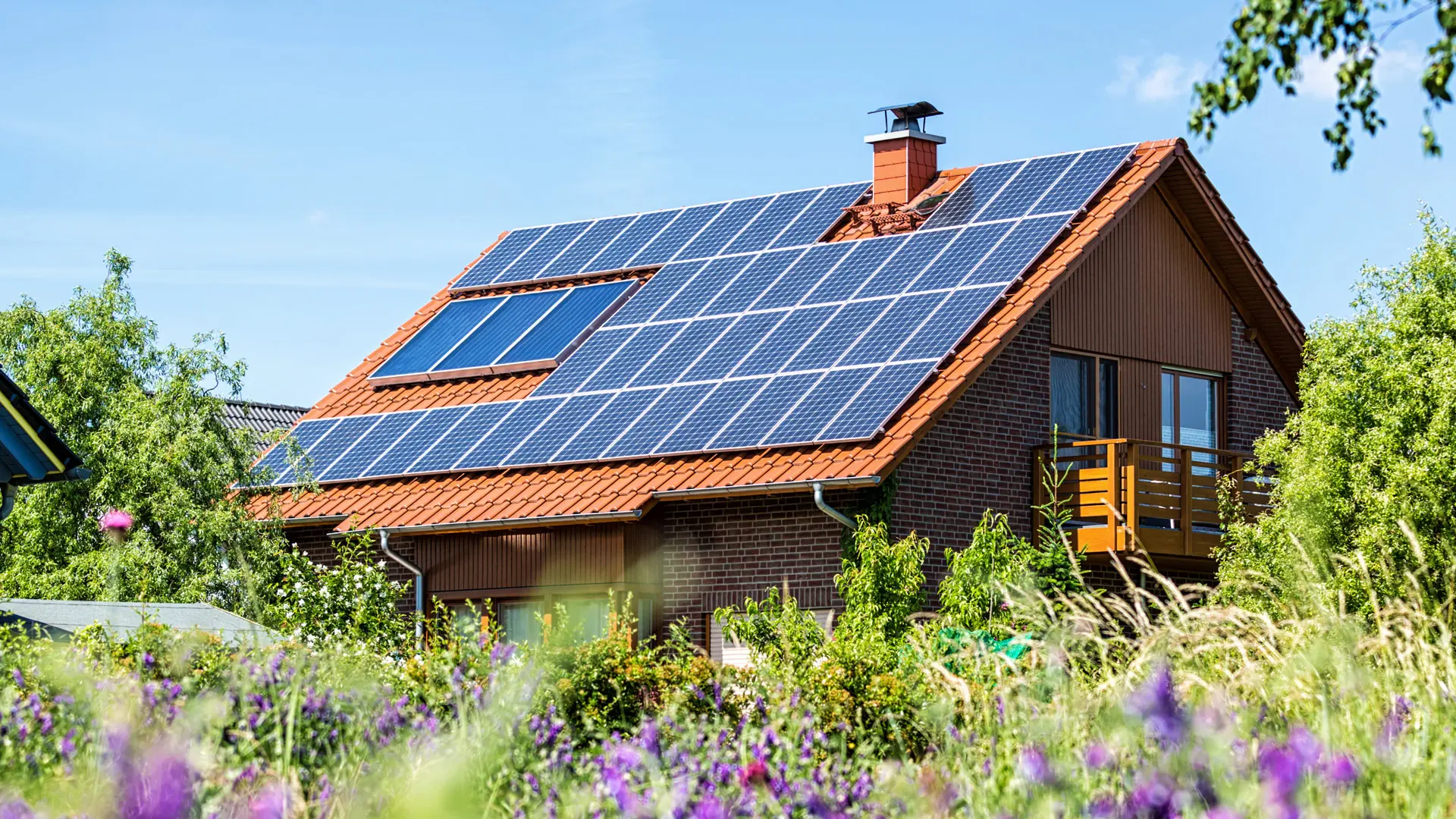 Haus mit Solarpanelen auf dem Dach, umgeben von grüner Vegetation und bunten Blumen im Vordergrund.