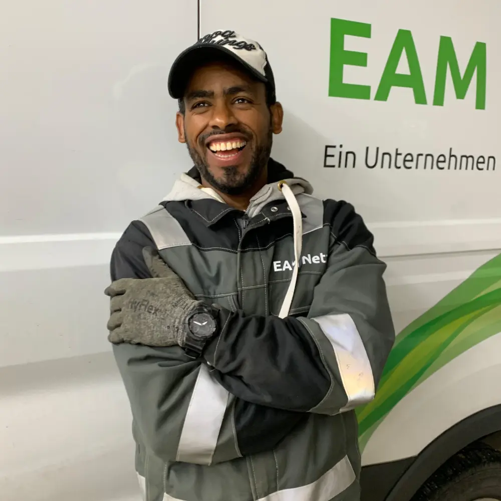 Lächelnder Mann in Arbeitskleidung mit Handschuhen steht vor einem Fahrzeug mit dem Schriftzug "EAM""."