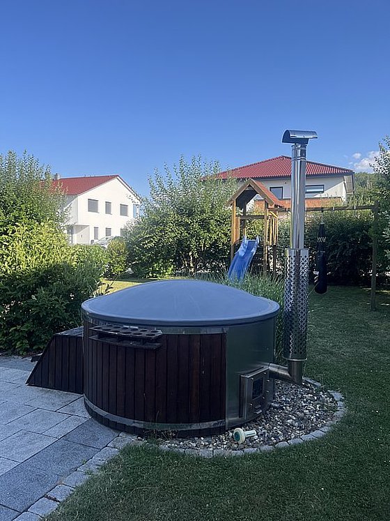 Holzofen mit runder Abdeckung im Garten, umgeben von Sträuchern und einem Spielplatz im Hintergrund. Klarer blauer Himmel.