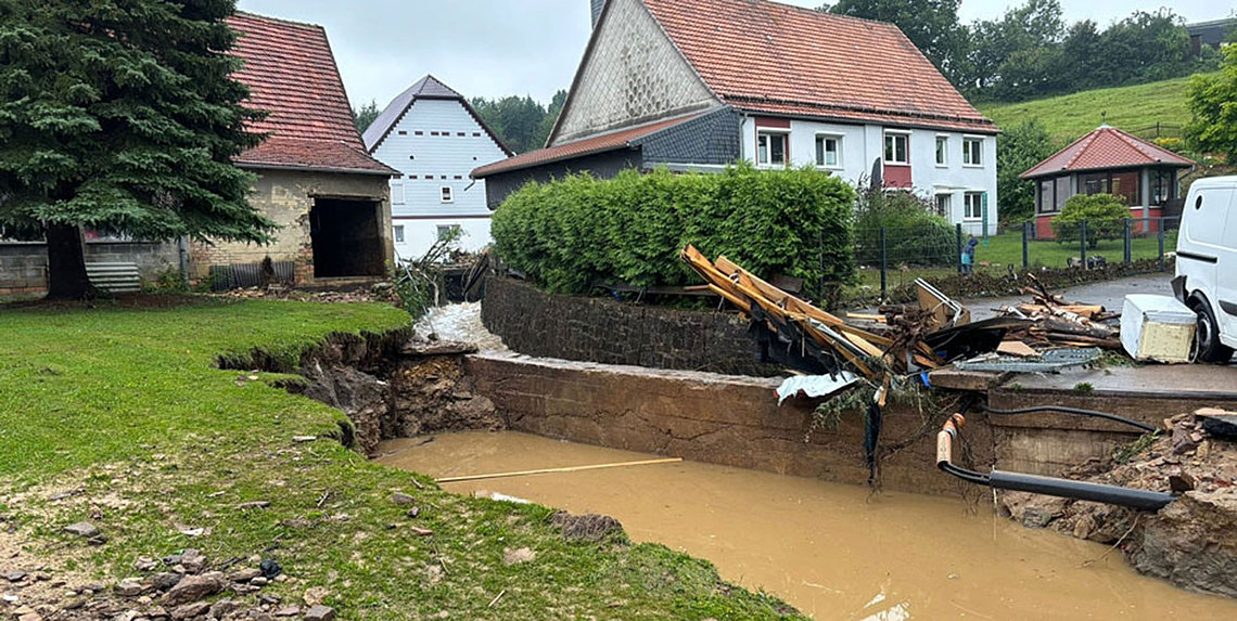 Nach einem Hochwasser fließt trübes Wasser durch eine Schlammgrube mit Trümmern, während links ein Tannenbaum und im Hintergrund Häuser stehen.