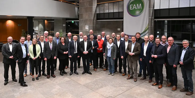 Eine große Gruppe von Personen posiert in einem modernen Innenraum, umgeben von Pflanzen und einem Banner im Hintergrund.