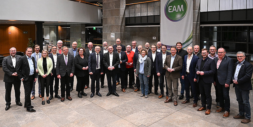 Eine große Gruppe von Personen posiert in einem modernen Innenraum, umgeben von Pflanzen und einem Banner im Hintergrund.