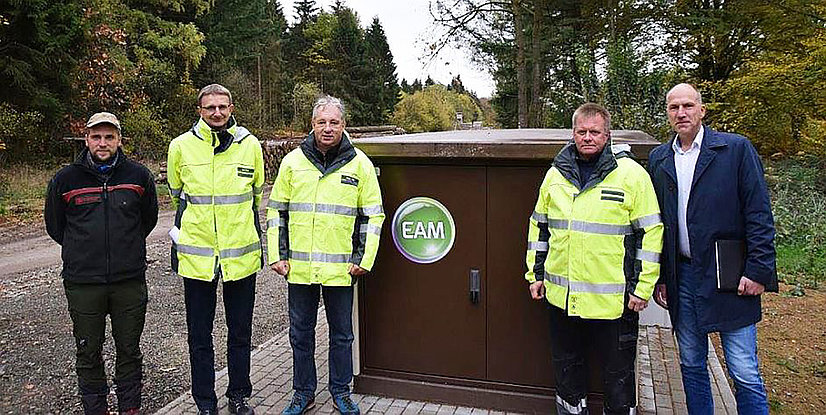 Fünf Personen in Sicherheitskleidung stehen vor einem Technikschrank im Wald, umgeben von Bäumen und herbstlichen Farben.