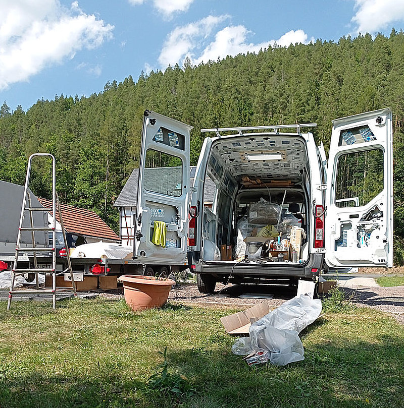 Ein offener Lieferwagen steht auf einer Wiese, umgeben von Bäumen und einem Haus, mit Werkzeugen und Materialien im Inneren.