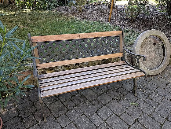 Eine Holzbänke mit einem metallenen Gestell in einem Garten, umgeben von Pflanzen und einem runden Stein.