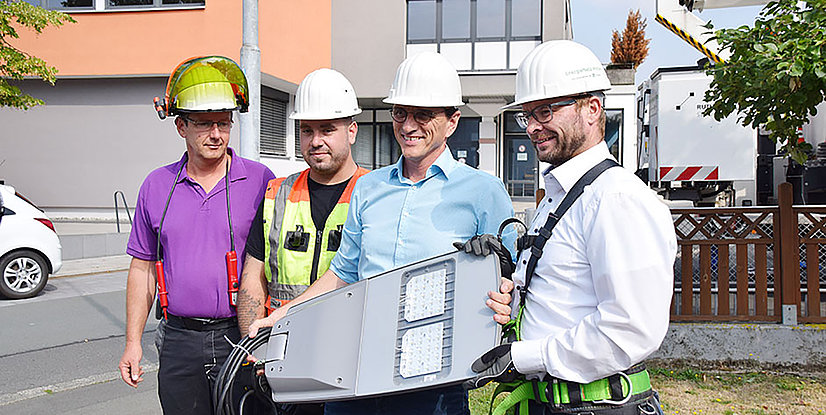 Vier Männer in Arbeitskleidung und Schutzhelm stehen draußen zusammen und halten ein neues Lichtgerät.