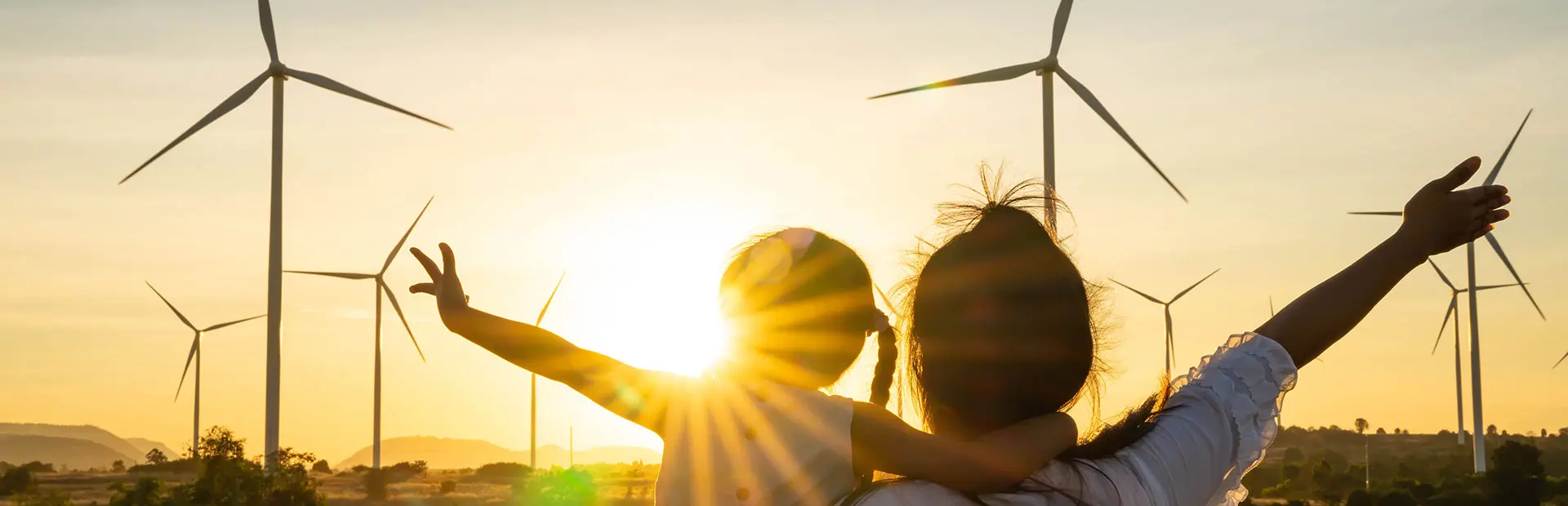 Mutter und Kind von hinten, Arme ausgestreckt, blicken bei Sonnenuntergang auf Windräder am Horizont.
