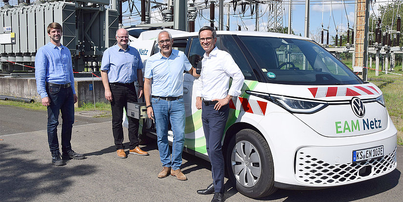 Vier Personen stehen neben einem Volkswagen ID. Buzz mit EAM Netz Beschriftung vor einem Stromnetz.