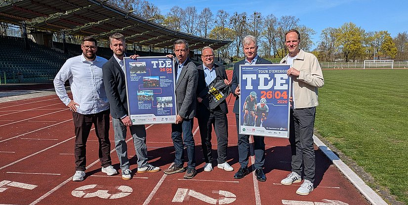 Sechs Personen stehen auf einer Laufbahn in einem Stadion und halten Veranstaltungsplakate in die Kamera, darunter ein Plakat zur „Tour d’Energie“ mit dem Datum 26.04.2026; im Hintergrund sind Tribüne, Rasenfläche und Bäume bei sonnigem Wetter zu sehen.