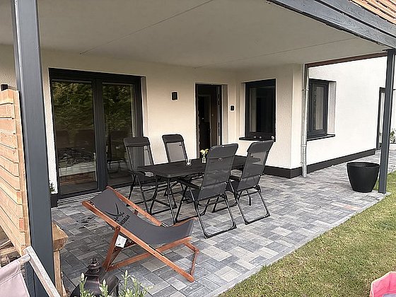 Moderne Terrasse mit einem Tisch, sechs Stühlen, einer Liege und einer Pflanze vor einem stilvollen Haus.