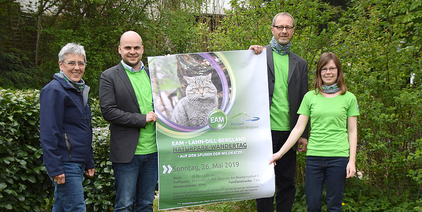 Vier Personen mit grünen T-Shirts halten ein Plakat für den Naturpark-Wandertag im Freien, umgeben von Pflanzen.