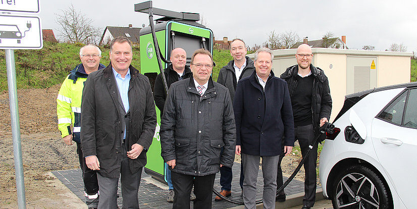 Gruppe von Personen an einer Elektro-Ladestation, hinter ihnen ein Elektroauto und ein Verkehrsschild für E-Ladestationen.