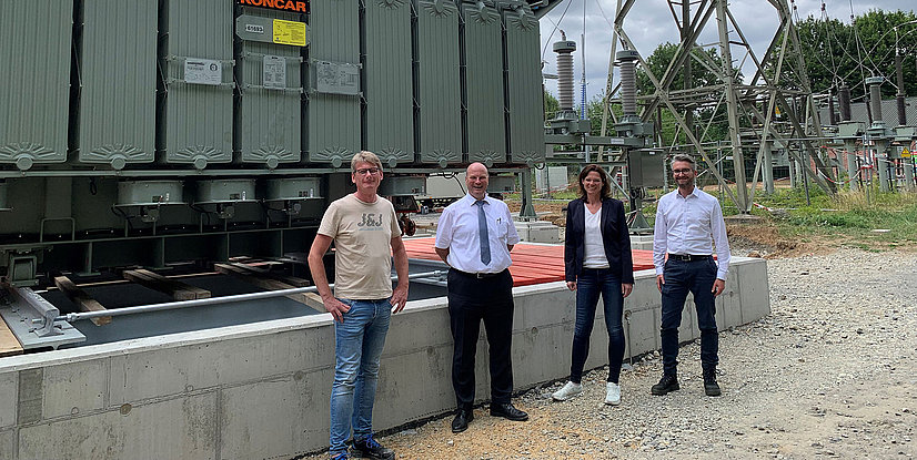 Vier Personen stehen vor einem Transformator in einem Umspannwerk, umgeben von elektrischen Anlagen und Freileitungen.