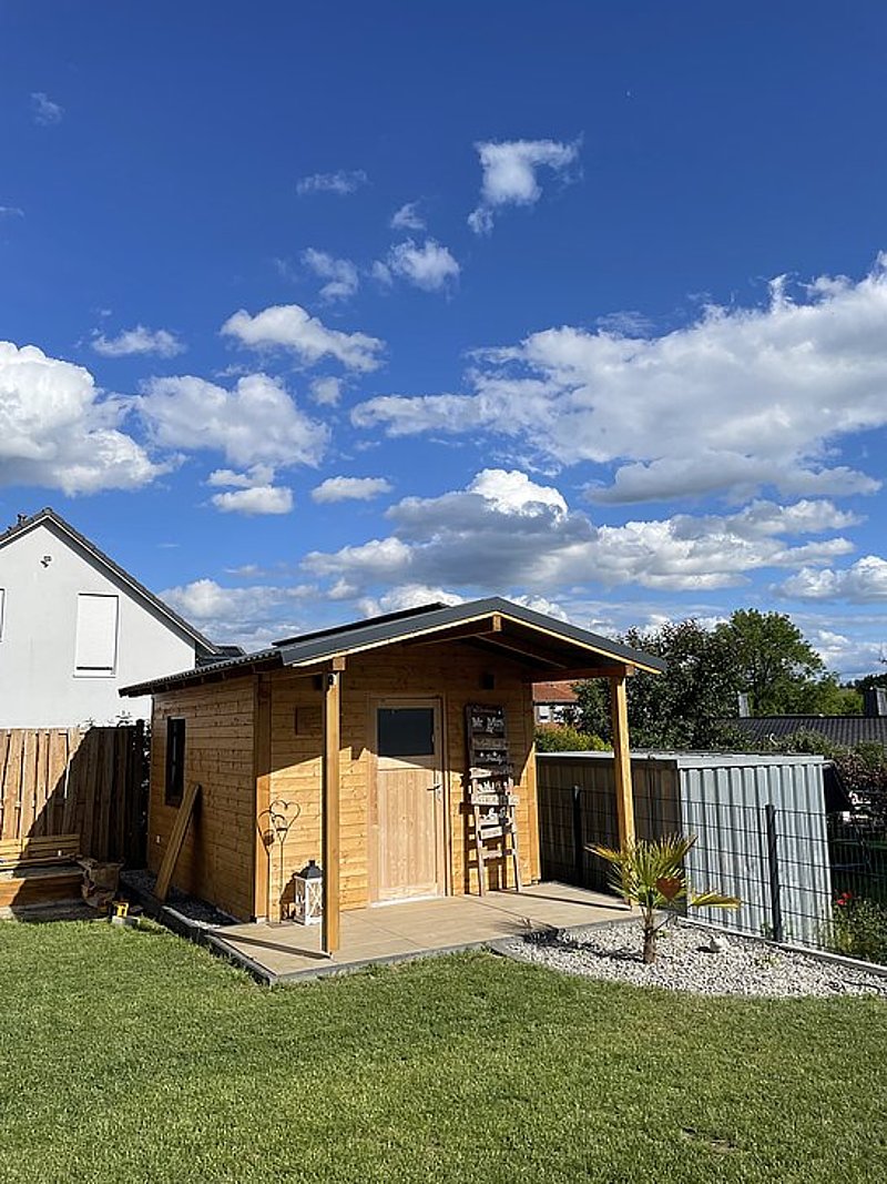 Holzhaus im Garten unter blauem Himmel mit Wolken, umgeben von Rasen und Zaun.