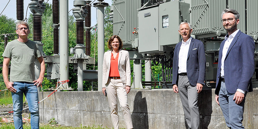 Vier Personen stehen vor einer elektrischen Anlage im Freien, umgeben von grüner Vegetation.