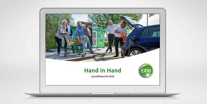 Ein Laptop zeigt ein Bild von Menschen, die vor einem Supermarkt einkaufen und Taschen in ein Auto laden. Darunter steht Hand in Hand und das EAM-Logo.