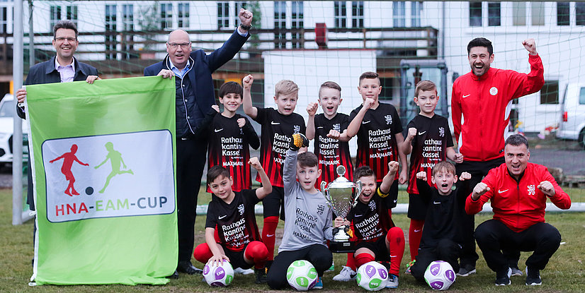 Jubelnde Kinderfußballmannschaft mit Pokal und Banner des HNA-EAM-Cups auf dem Spielfeld.