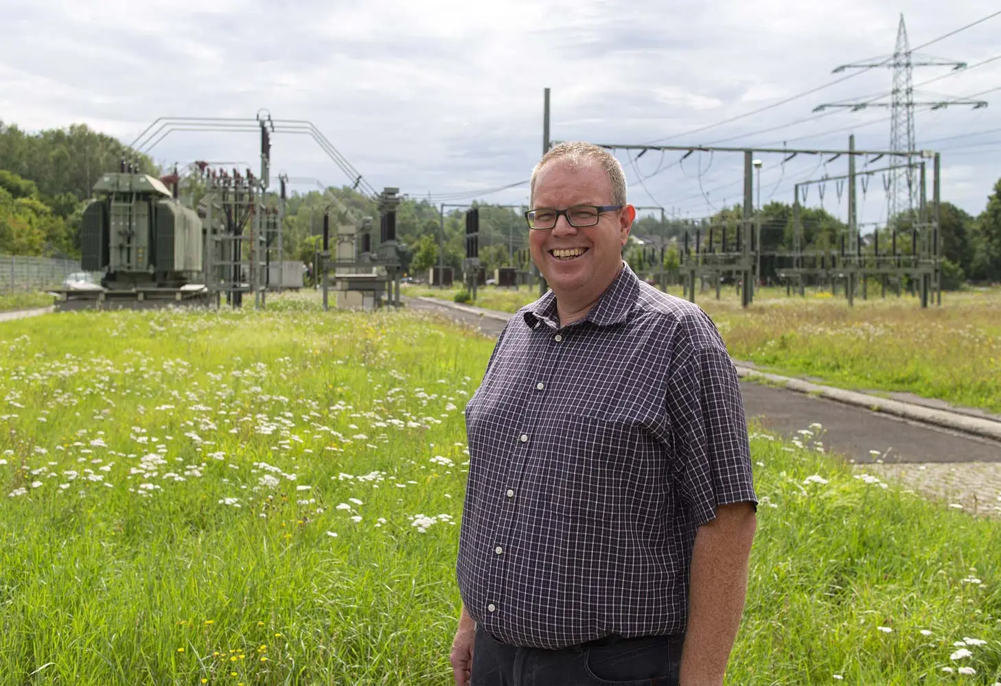 Mann mit Brille und kariertem Hemd steht auf einer Wiese mit Blumen, im Hintergrund sind Strommasten und Transformatoren sichtbar.