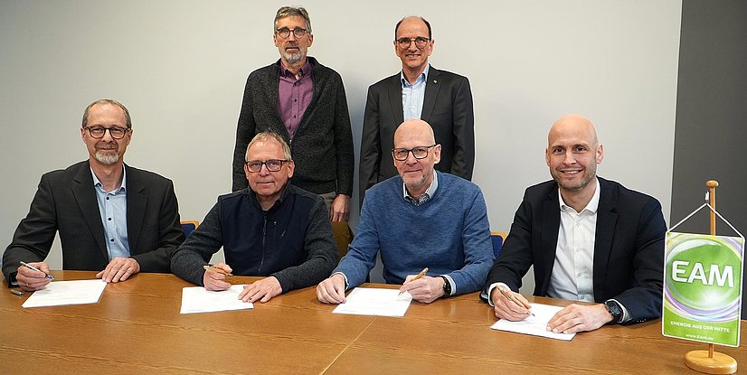 Sieben Personen sind an einem Tisch in einem Besprechungsraum versammelt; vier Personen sitzen und unterzeichnen Dokumente, drei weitere stehen dahinter. Auf dem Tisch steht eine kleine Tischfahne mit dem Schriftzug „EAM“.