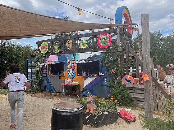 Bild eines bunten Außencafés mit einem künstlerisch gestalteten Container, dekorativen Elementen und einer Person im Hintergrund.