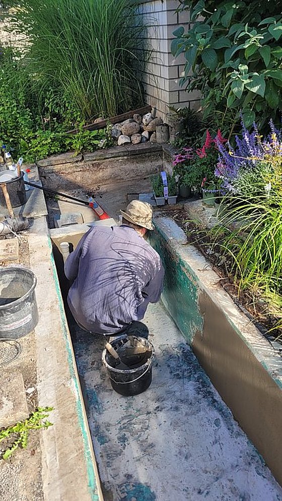 Ein Arbeiter, der in einem bepflanzten Garten einen Graben gräbt, umgeben von Pflanzen und Werkzeugen.