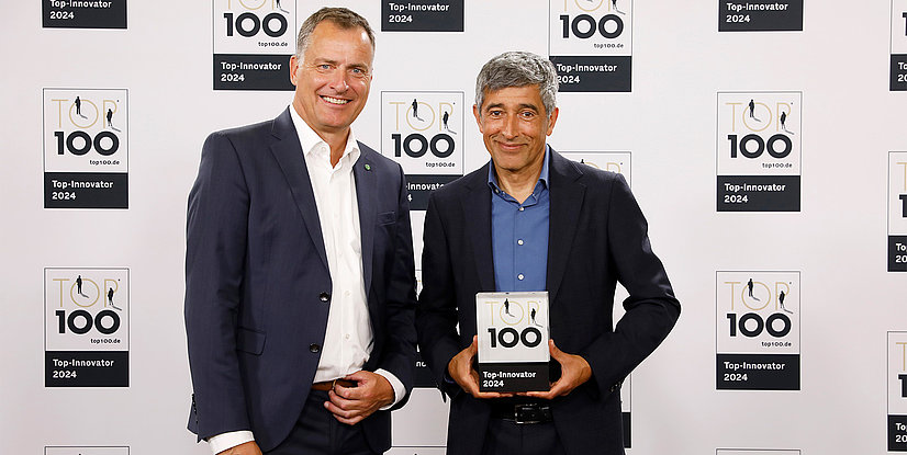 Zwei Männer lächeln und halten eine Auszeichnung in der Hand, vor einem Hintergrund mit Logos von Top 100 - Top-Innovator 2024.