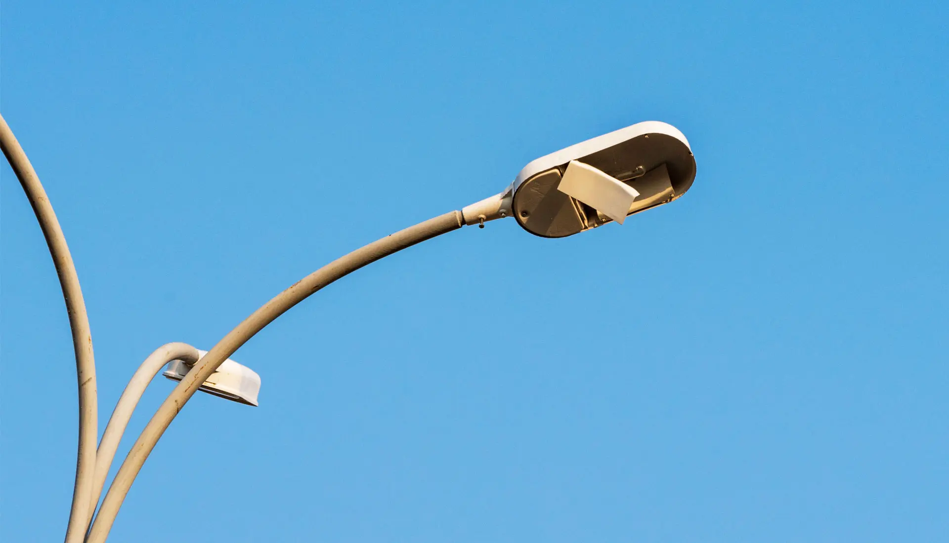 Gebogener Arm einer Straßenlaterne mit defektem, leerem Lampengehäuse vor blauem Himmel.