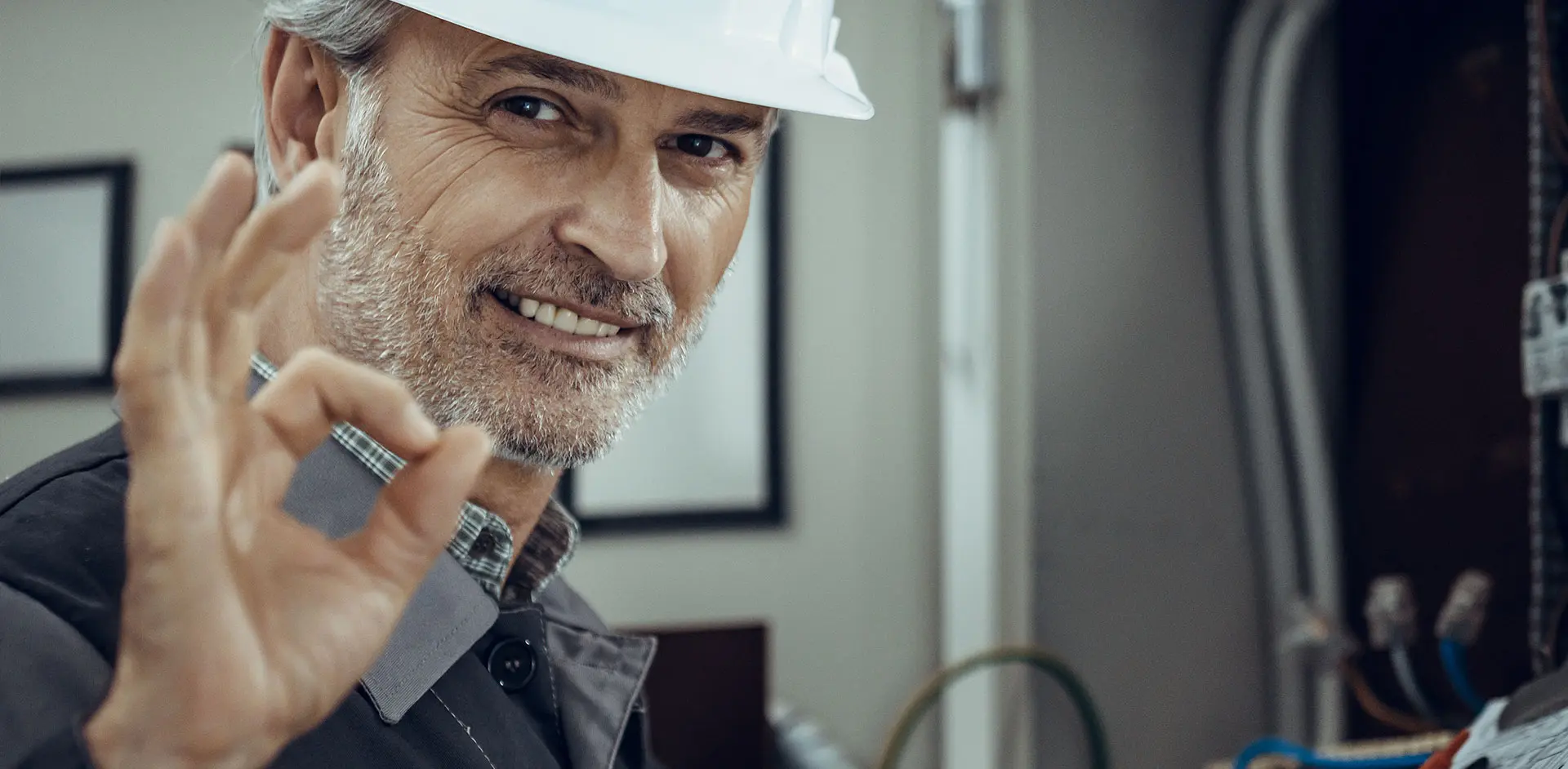  Lächelnder Ingenieur mit Schutzhelm und grauem Bart zeigt das "Okay"-Zeichen, während er in die Kamera blickt.