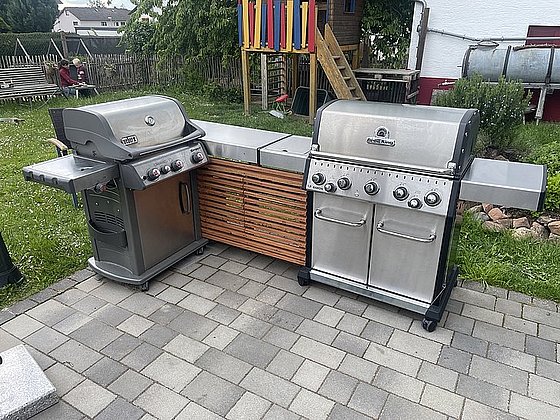 Zwei platzierte Grills auf einem gepflasterten Boden, umgeben von Gartenmöbeln und Spielgeräten.