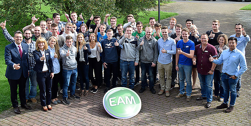 Gruppenshot von Menschen, die zusammen stehen und Daumen hoch zeigen, vor einer grünen Wiese mit dem EAM-Logo im Vordergrund.