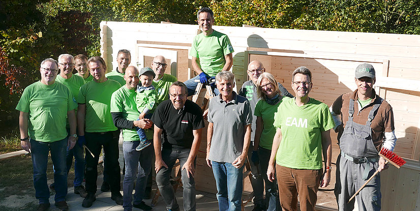 Gruppe von Menschen in grünen T-Shirts, die vor einem Holzbau stehen, lächeln und posieren zusammen im Freien.
